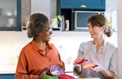Happy caregiver and senior talking at home. Elderly female and nurse are with vegetables. They are preparing food in kitchen.