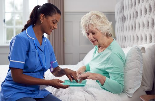 Female Care Worker In Uniform Helping Senior Woman At Home In Bed With Medication